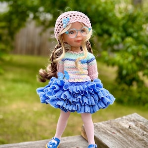 May include: A doll wearing a pink crocheted hat, round glasses, and a colorful sweater with a blue ruffled skirt. The doll has blonde hair and is wearing light pink tights and blue shoes with yellow soles. The background is a blurred green.