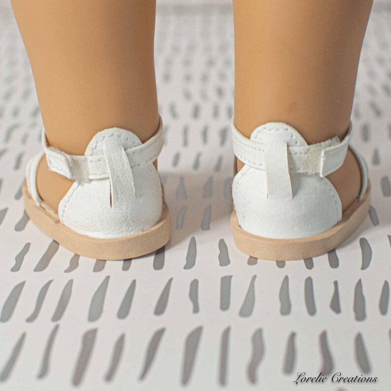 Puede incluir: Primer plano de dos sandalias blancas para mu&ntilde;eca, con suelas beige y correas de tobillo ajustables. Las sandalias tienen un dise&ntilde;o de gladiador, con tal&oacute;n cerrado y parte delantera abierta. El fondo es una superficie blanca con un patr&oacute;n gris.