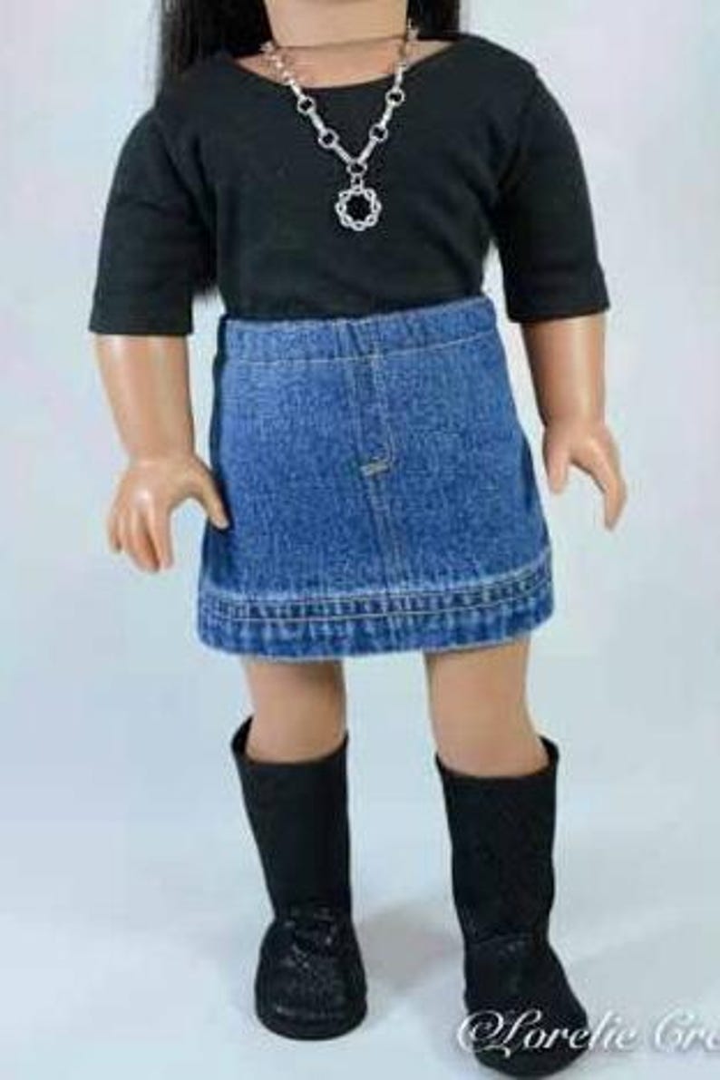Puede incluir: Una mu&ntilde;eca con una camisa negra de manga larga, una falda vaquera y botas negras. La mu&ntilde;eca lleva un collar de plata con un colgante circular.