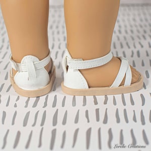 Puede incluir: Sandalias blancas para mu&ntilde;eca, con suela marr&oacute;n claro y correas blancas. Las sandalias tienen un dise&ntilde;o sencillo con una correa alrededor del tobillo y sobre el pie. Las sandalias est&aacute;n dise&ntilde;adas para una mu&ntilde;eca.