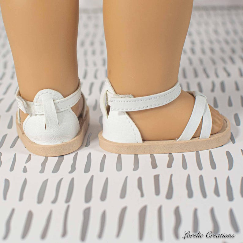 Puede incluir: Sandalias blancas para mu&ntilde;eca, con suela marr&oacute;n claro y correas blancas. Las sandalias tienen un dise&ntilde;o sencillo con una correa alrededor del tobillo y sobre el pie. Las sandalias est&aacute;n dise&ntilde;adas para una mu&ntilde;eca.
