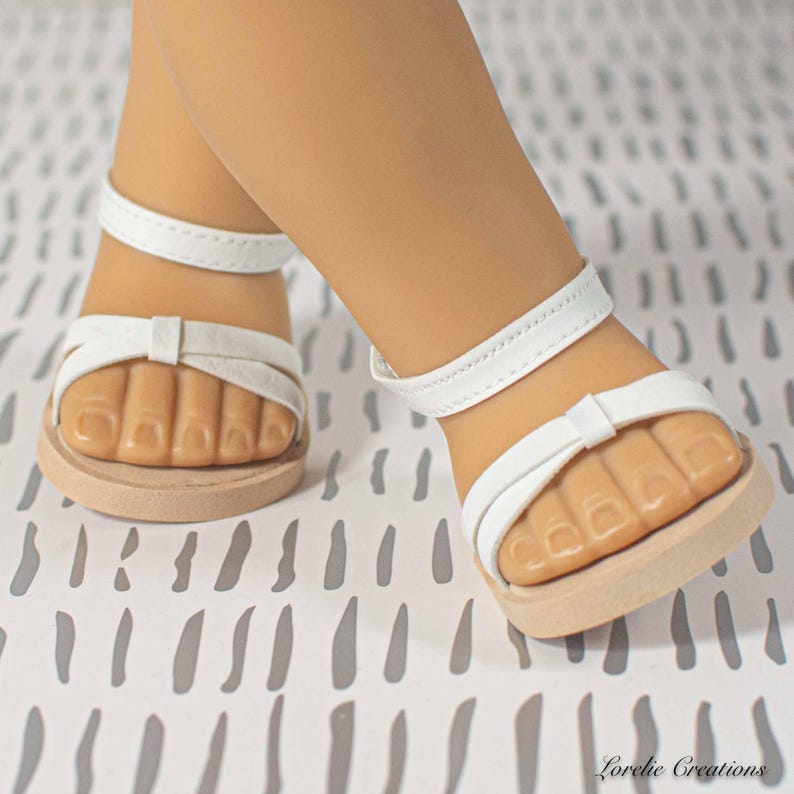 Puede incluir: Sandalias blancas para mu&ntilde;eca con un lazo en la correa delantera y una correa en el tobillo. Las sandalias tienen una suela marr&oacute;n claro. Las sandalias est&aacute;n en los pies de una mu&ntilde;eca.