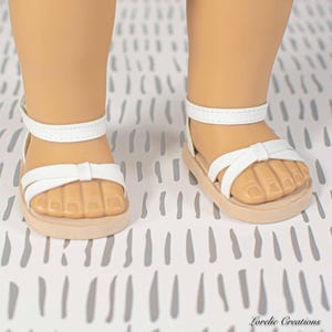 Puede incluir: Sandalias blancas con detalle de lazo y correa en el tobillo. Las sandalias tienen suela marr&oacute;n claro y est&aacute;n dise&ntilde;adas para una mu&ntilde;eca. Las sandalias se muestran sobre una superficie estampada.