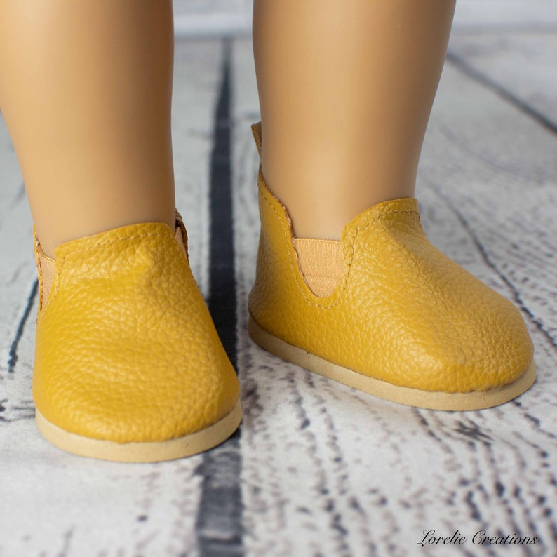 Puede incluir: Un par de zapatos amarillos de vestir para una mu&ntilde;eca. Los zapatos est&aacute;n hechos de un material texturizado similar al cuero y tienen una suela marr&oacute;n claro.