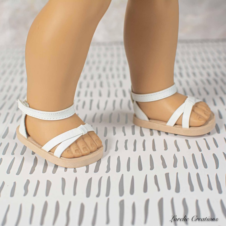 Puede incluir: Sandalias blancas con suela marr&oacute;n claro. Las sandalias tienen una correa en el tobillo y correas en la parte superior del pie. Estas sandalias est&aacute;n dise&ntilde;adas para una mu&ntilde;eca.