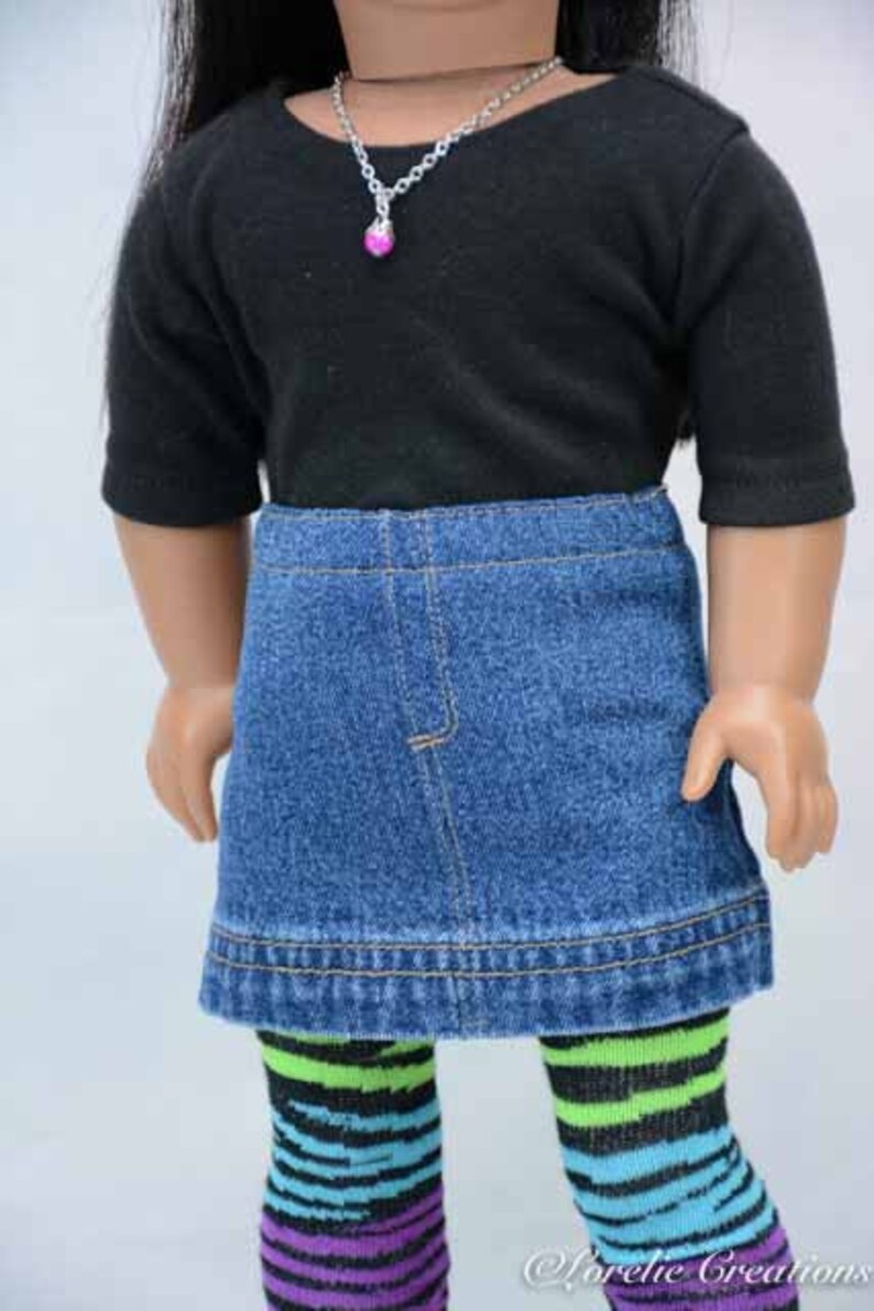 Puede incluir: Una mu&ntilde;eca con una camisa negra de manga corta, una falda vaquera y medias de rayas de colores. La mu&ntilde;eca lleva un collar de cadena plateada con un colgante de coraz&oacute;n rosa.