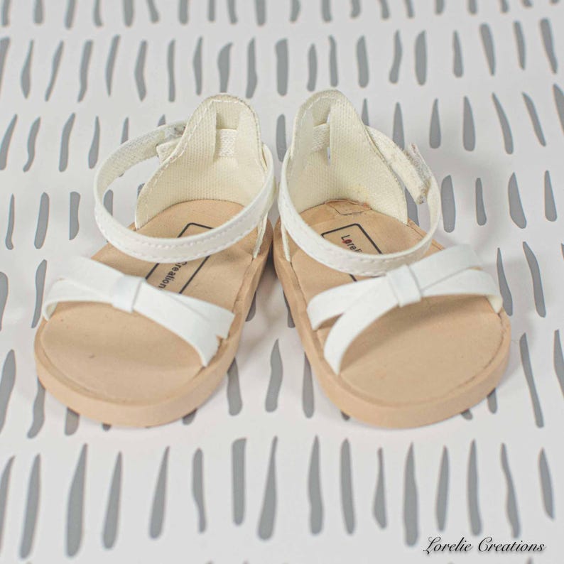 Puede incluir: Un par de sandalias blancas con un detalle de lazo en las correas delanteras. Las sandalias tienen una suela beige y correas de tobillo blancas con hebillas ajustables. Las sandalias est&aacute;n dise&ntilde;adas para una mu&ntilde;eca o un ni&ntilde;o peque&ntilde;o.