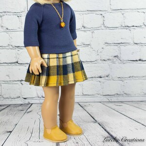 Puede incluir: Una mu&ntilde;eca con una camisa de manga larga azul marino con un collar de cadena dorada y una falda de cuadros amarillos y azules. La mu&ntilde;eca lleva zapatos marrones.