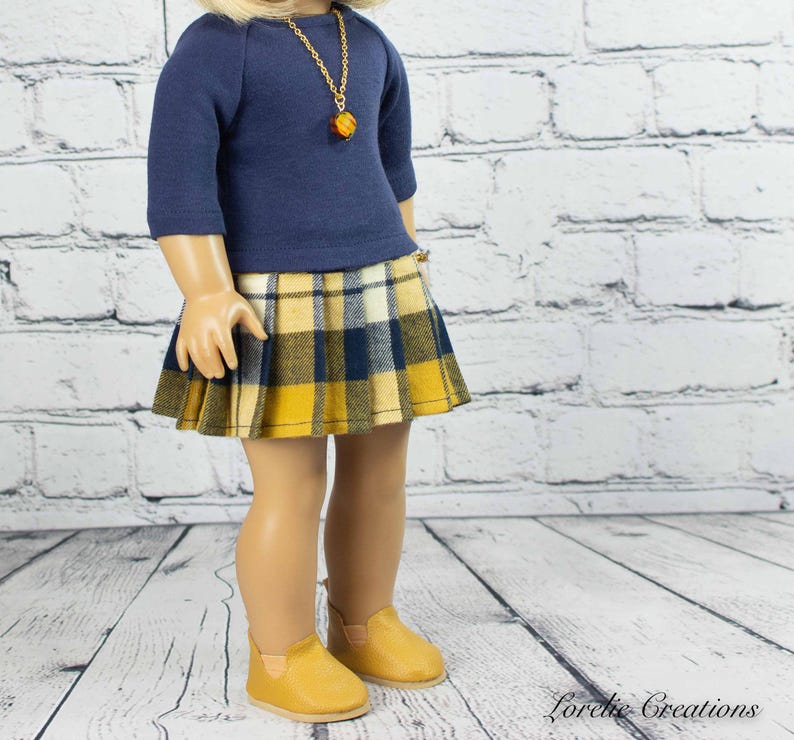 Puede incluir: Una mu&ntilde;eca con una camisa de manga larga azul marino con un collar de cadena dorada y una falda de cuadros amarillos y azules. La mu&ntilde;eca lleva zapatos marrones.