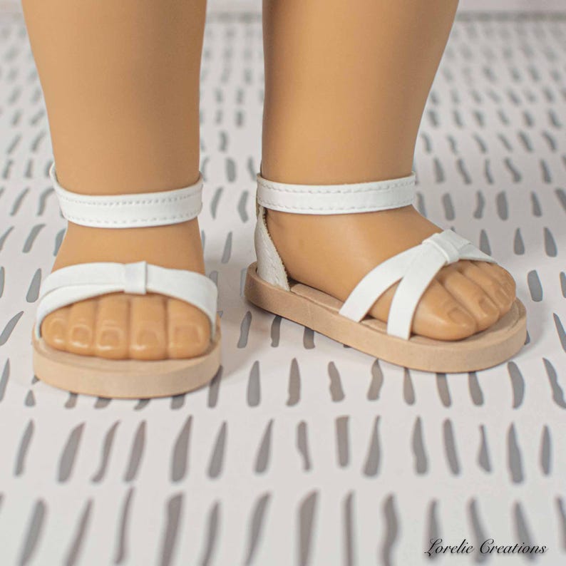 Puede incluir: Sandalias blancas con detalle de lazo y correa en el tobillo. Las sandalias tienen una suela marr&oacute;n claro y est&aacute;n dise&ntilde;adas para una mu&ntilde;eca. Las sandalias se muestran en los pies de una mu&ntilde;eca.