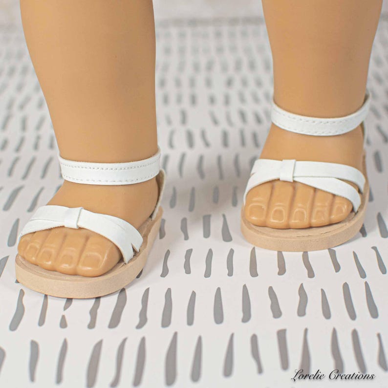 Puede incluir: Sandalias blancas con detalle de lazo y correa en el tobillo. Las sandalias tienen una suela marr&oacute;n claro y est&aacute;n dise&ntilde;adas para una mu&ntilde;eca. Las sandalias se muestran sobre una superficie con estampado blanco y gris.