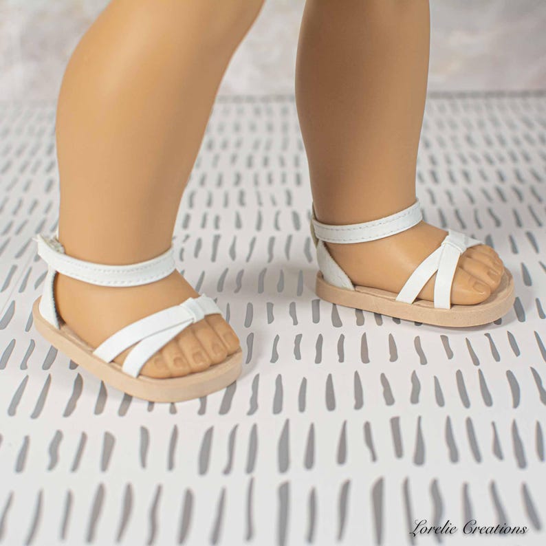 Puede incluir: Sandalias blancas con suela marr&oacute;n claro. Las sandalias tienen una correa en el tobillo y un dise&ntilde;o entrecruzado sobre el pie. Las sandalias est&aacute;n dise&ntilde;adas para una mu&ntilde;eca.