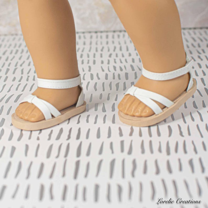 Puede incluir: Sandalias blancas con suela marr&oacute;n claro. Las sandalias tienen una correa alrededor del tobillo y un dise&ntilde;o entrecruzado sobre el pie. Estas sandalias son para una mu&ntilde;eca.