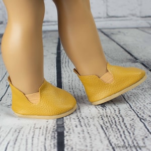 Puede incluir: Un par de zapatos amarillos mostaza para una mu&ntilde;eca. Los zapatos tienen un acabado texturizado similar al cuero y una punta ligeramente redondeada.