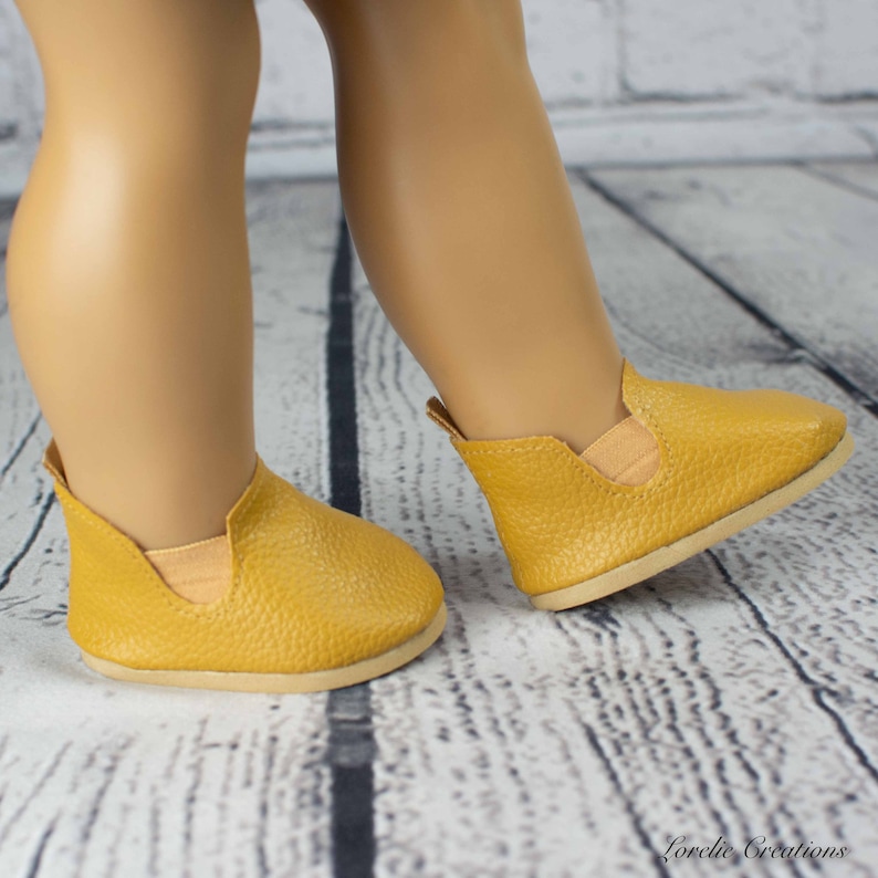 Puede incluir: Un par de zapatos amarillos mostaza para una mu&ntilde;eca. Los zapatos tienen un acabado texturizado similar al cuero y una punta ligeramente redondeada.