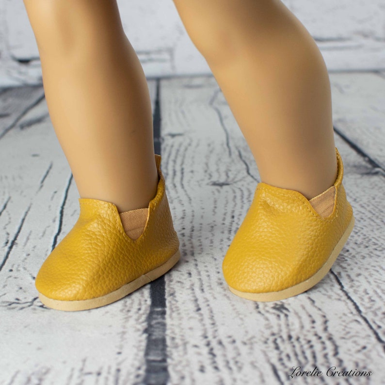 Puede incluir: Un par de zapatos amarillos mostaza para una mu&ntilde;eca. Los zapatos tienen un acabado texturizado similar al cuero y una punta ligeramente redondeada.