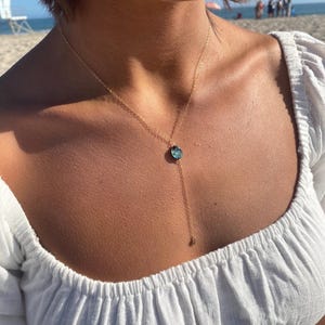 May include: A gold chain necklace with a blue gemstone pendant. The necklace is worn by a person with a white top.