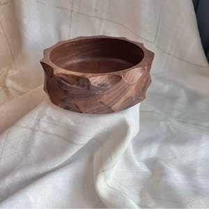May include: A hand-carved wooden bowl with a faceted exterior. The bowl is a rich brown color and has a smooth interior. The bowl is sitting on a white fabric.