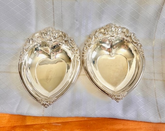 May include: Two silver heart-shaped dishes with ornate detailing. The dishes are made of polished silver and have a heart-shaped indentation in the center.