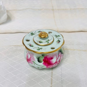 May include: A small, round, porcelain trinket box with a lid. The box is decorated with a floral pattern of pink roses and green leaves on a light blue background. The lid has a gold knob and a gold trim around the edge.