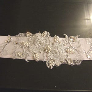 May include: A white satin bridal sash with silver floral embroidery and rhinestones.