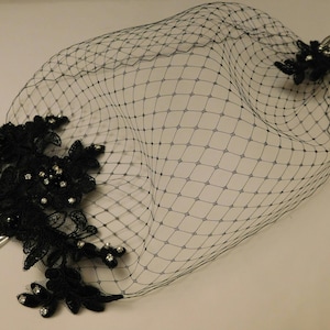 black birdcage veil with a delicate floral lace design and a comb for attaching to hair. The veil is made of fine netting and has a soft, flowing shape.