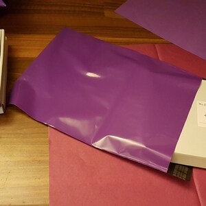 May include: Two white boxes with the text "The Dragon - Bead Alchemy" and a black and white illustration of a dragon. The boxes are wrapped in a purple poly mailer.