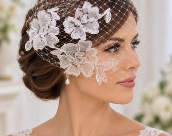 May include: A white birdcage veil featuring a fine diamond-patterned netting adorned with three distinct white floral lace appliqués. The veil is positioned over the side of the head, with the lace flowers layered across the netting.