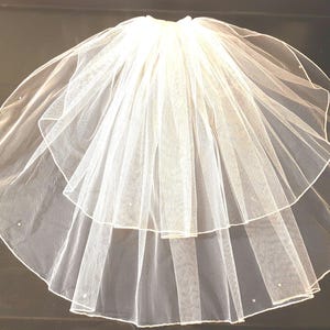 Puede incluir: Velo de novia de tul blanco de dos niveles. El velo está recogido en la parte superior y se ensancha, con una apariencia delicada y transparente. Pequeños adornos dispersos añaden un brillo sutil. Ideal para bodas.