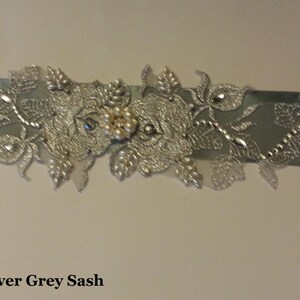 May include: A silver grey sash with a silver floral design and pearl embellishments. The sash is decorated with a large silver flower with a cluster of pearls in the center.