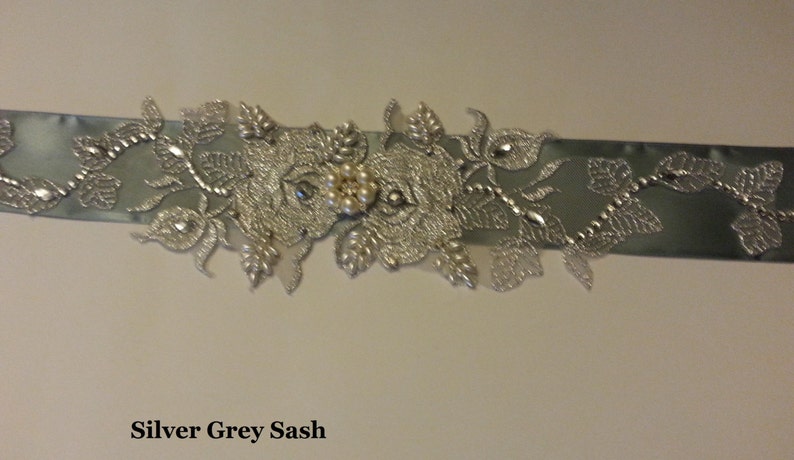 May include: A silver grey sash with a silver floral design and pearl embellishments. The sash is decorated with a large silver flower with a cluster of pearls in the center.