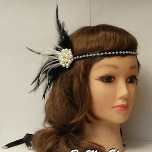 Puede incluir: Una diadema negra con una fila de pedrería y un adorno de plumas blancas y negras con un adorno de perlas. La diadema se lleva en una cabeza de maniquí con pelo castaño.