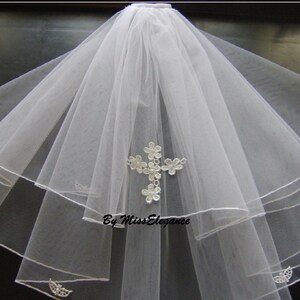 May include: White tulle veil with a cross design made of white lace flowers. The veil has three layers and is trimmed with white satin.
