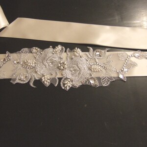 May include: A white satin ribbon with a silver floral embellishment featuring two large roses and leaves. The embellishment is adorned with rhinestones and pearls.