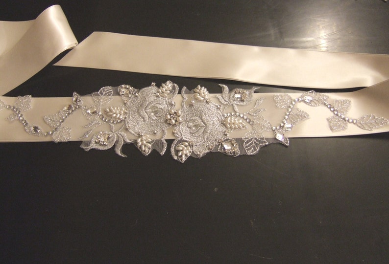 May include: A white satin ribbon with a silver floral embellishment featuring two large roses and leaves. The embellishment is adorned with rhinestones and pearls.