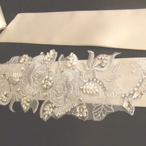 May include: A white satin bridal sash with a silver floral embellishment featuring pearls and rhinestones.
