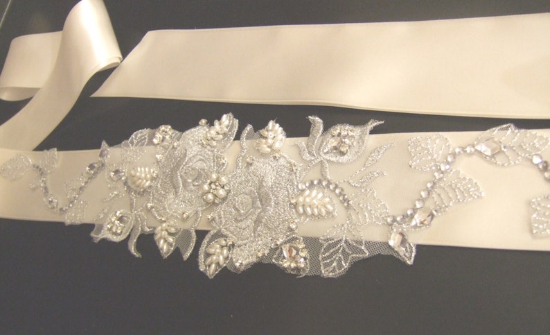May include: A white satin bridal sash with a silver floral embellishment featuring pearls and rhinestones.