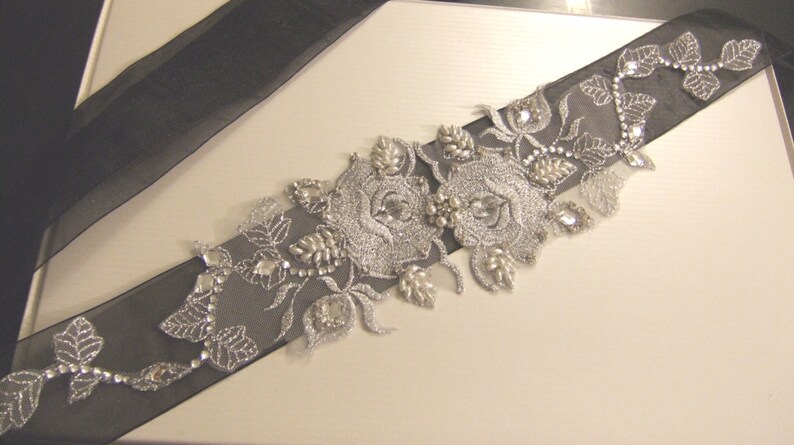 May include: A black satin ribbon with silver embroidery and rhinestones, featuring a floral design with two large roses. The ribbon is perfect for adding a touch of elegance to any occasion.