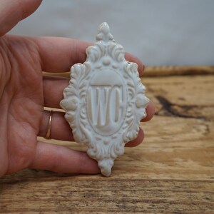 May include: White ceramic sign with an ornate design and the letters "WC" in the center.