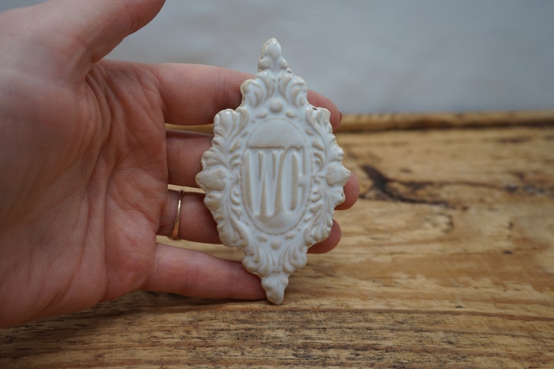 May include: White ceramic sign with an ornate design and the letters "WC" in the center.