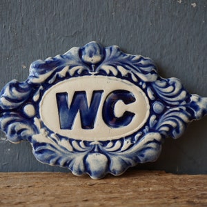 May include: A blue and white ceramic sign with a decorative border and the letters "WC" in blue.