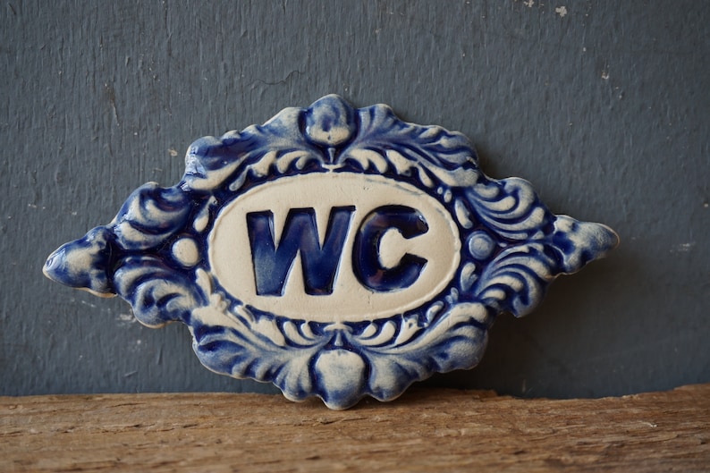 May include: A blue and white ceramic sign with a decorative border and the letters "WC" in blue.