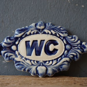 May include: A blue and white ceramic sign with a decorative border and the letters "WC" in blue.