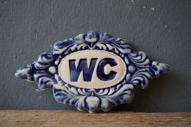 May include: A blue and white ceramic sign with a decorative border and the letters "WC" in blue.