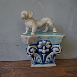 May include: A white ceramic dog figurine standing on a blue and white ceramic pedestal with intricate floral designs.