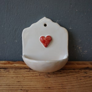 May include: A white ceramic wall pocket with a scalloped top and a red heart in the centre. The pocket has a small, rounded basin at the bottom and a hole at the top for hanging. The background is a dark blue-grey.