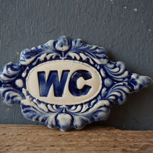 May include: A blue and white ceramic sign with the letters WC in a white oval surrounded by a blue and white floral design.