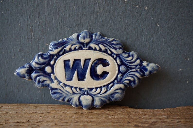May include: A blue and white ceramic sign with the letters WC in a white oval surrounded by a blue and white floral design.