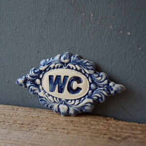 May include: A blue and white ceramic sign with a decorative border and the letters "WC" in a white oval.
