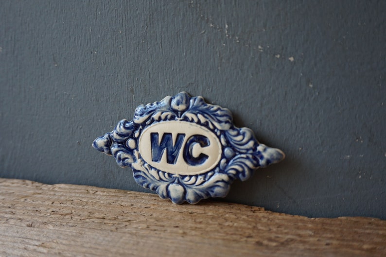 May include: A blue and white ceramic sign with a decorative border and the letters "WC" in a white oval.